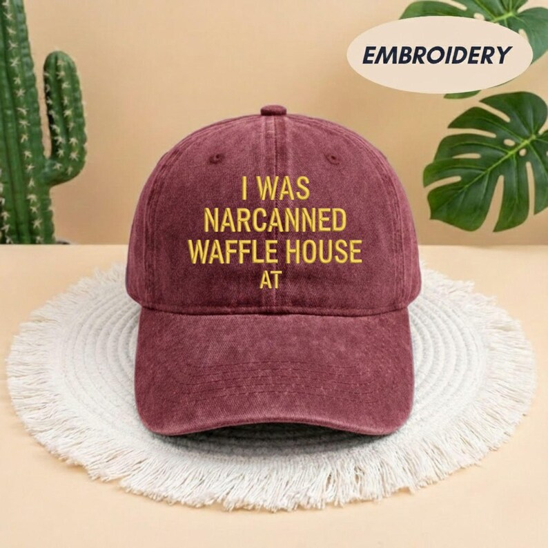May include: A maroon baseball cap with the embroidered text "I WAS NARCANNED WAFFLE HOUSE AT" in yellow. The hat is displayed on a white, fringed placemat. The word "EMBROIDERY" is printed on a white oval.