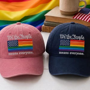 May include: Two baseball caps, one dusty rose and one navy blue, each featuring embroidered text that reads We the People and means everyone. Below the text is an embroidered American flag with rainbow-colored stripes instead of red.