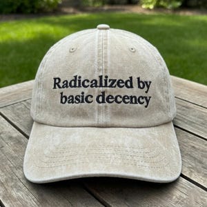 May include: A beige baseball cap with the embroidered text "Radicalized by basic decency" in black. The cap has a curved brim and a six-panel design. The hat is made of a soft, textured fabric.