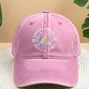 May include: A pink baseball cap with a curved peak. The cap has an embroidered circular design with a bird, flowers, and the text "RADICALIZED BY BASIC DECENCY".
