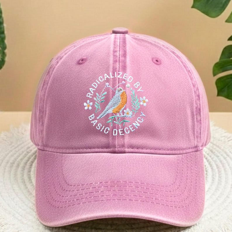 May include: A pink baseball cap with a curved peak. The cap has an embroidered circular design with a bird, flowers, and the text "RADICALIZED BY BASIC DECENCY".