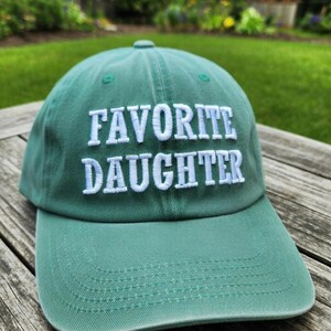 May include: A green baseball cap with the embroidered text "FAVORITE DAUGHTER" in white. The cap has a curved brim and a button on top. The hat is made of a soft fabric and is likely adjustable.
