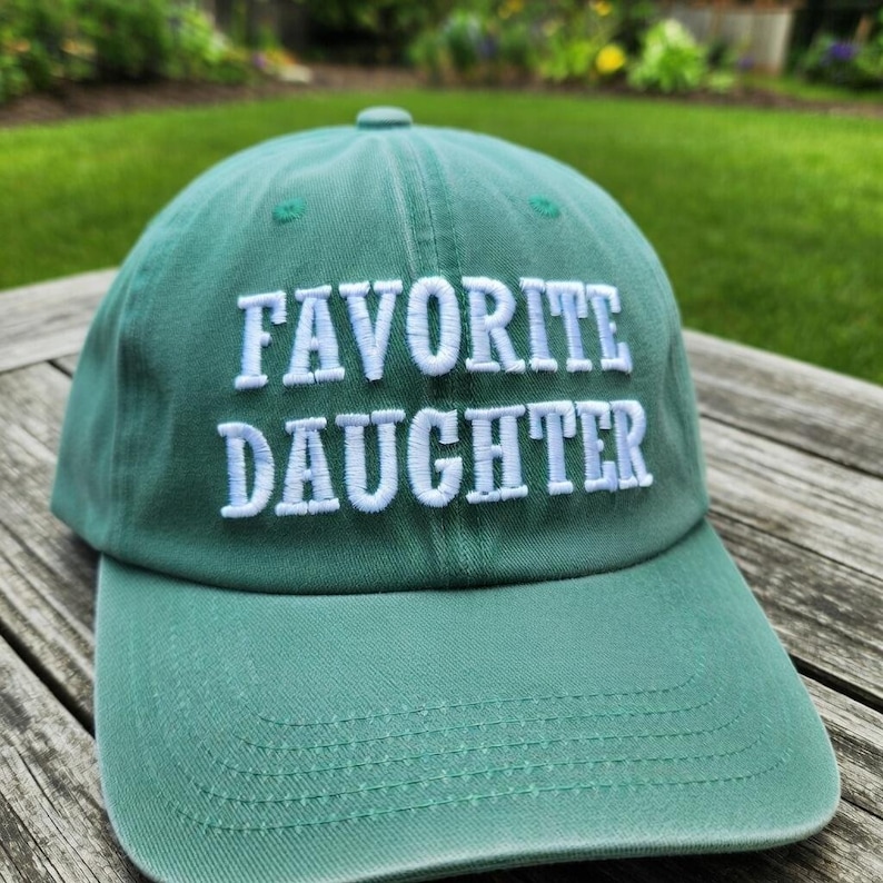 May include: A green baseball cap with the embroidered text "FAVORITE DAUGHTER" in white. The cap has a curved brim and a button on top. The hat is made of a soft fabric and is likely adjustable.