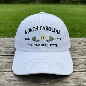 May include: White baseball cap with embroidered text and floral design. The text reads "NORTH CAROLINA EST. 1789 THE TAR HEEL STATE". The floral design features a white flower with a yellow centre and green leaves.
