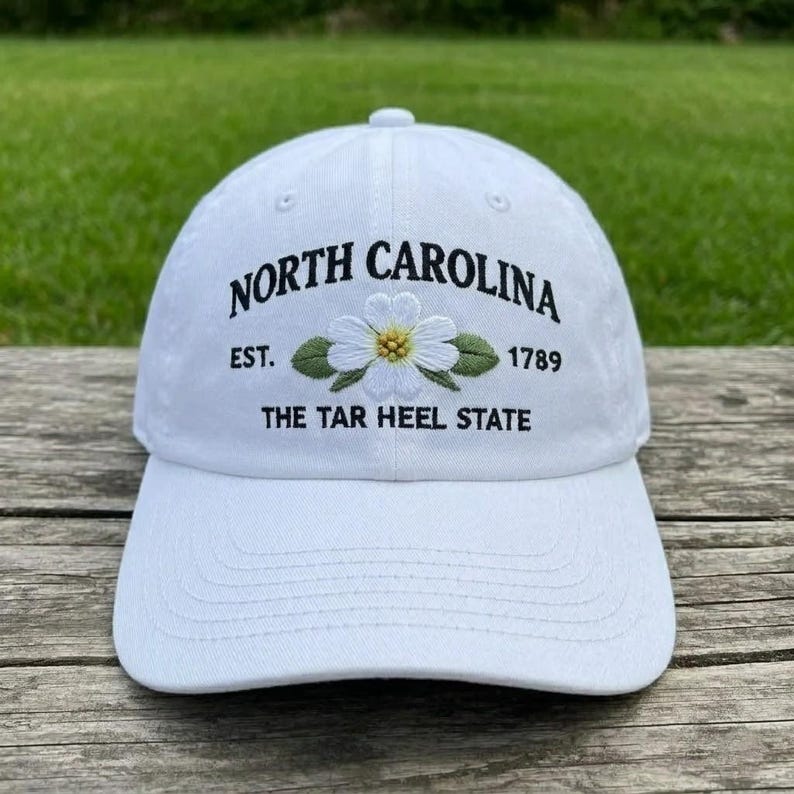 May include: White baseball cap with embroidered text and floral design. The text reads "NORTH CAROLINA EST. 1789 THE TAR HEEL STATE". The floral design features a white flower with a yellow centre and green leaves.