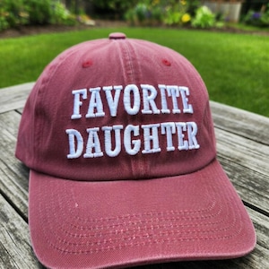 May include: A maroon baseball cap with the embroidered text "FAVORITE DAUGHTER" in white. The cap has a curved brim and a rounded top. The background is a blurred outdoor scene.