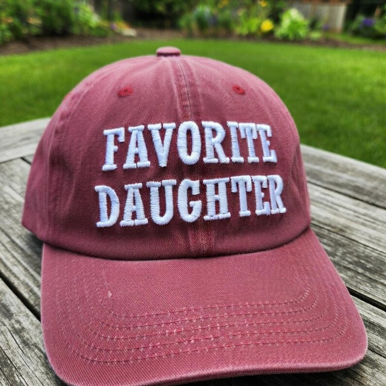 May include: A maroon baseball cap with the embroidered text "FAVORITE DAUGHTER" in white. The cap has a curved brim and a rounded top. The background is a blurred outdoor scene.
