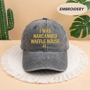 May include: Gray baseball cap with the embroidered text "I WAS NARCANNNED WAFFLE HOUSE AT" in yellow. The cap is sitting on a white, fringed placemat.