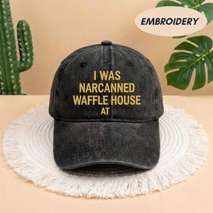 May include: Black baseball cap with the embroidered text "I WAS NARCANNED WAFFLE HOUSE AT" in yellow. The cap is made of a worn-looking fabric and has a curved brim. The word "EMBROIDERY" is in a white oval in the top right corner.