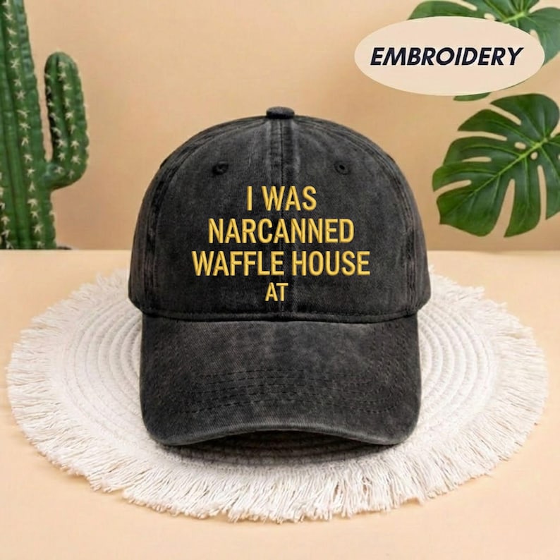 May include: Black baseball cap with the embroidered text "I WAS NARCANNED WAFFLE HOUSE AT" in yellow. The cap is made of a worn-looking fabric and has a curved brim. The word "EMBROIDERY" is in a white oval in the top right corner.
