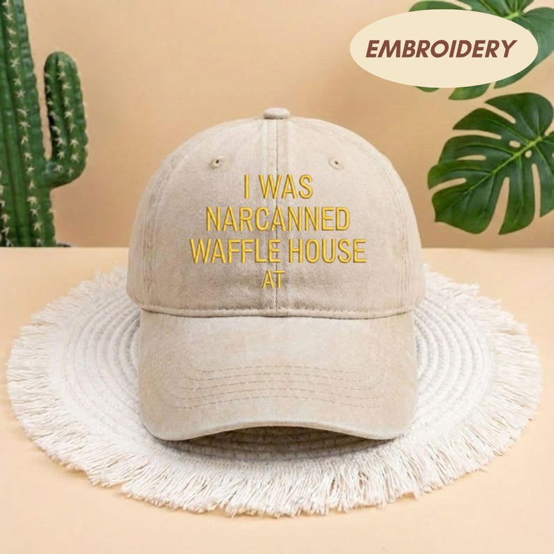 May include: A beige baseball cap with the embroidered text "I WAS NARCANNED WAFFLE HOUSE AT" in yellow. The cap is sitting on a white, fringed mat. The word "EMBROIDERY" is in a beige oval in the upper right corner.