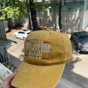 May include: Mustard-colored baseball cap with the text "MAKE AMERICA TRUMPLESS AGAIN" embroidered in white and red. The cap has a curved brim and a classic six-panel design. The hat is made of a soft, textured fabric.