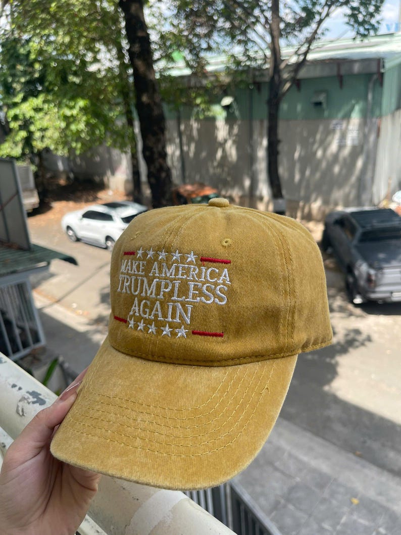 May include: Mustard-colored baseball cap with the text "MAKE AMERICA TRUMPLESS AGAIN" embroidered in white and red. The cap has a curved brim and a classic six-panel design. The hat is made of a soft, textured fabric.