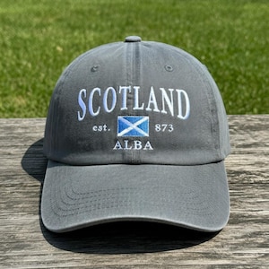 May include: Gray baseball cap with the word "SCOTLAND" embroidered in white, along with the Scottish flag and the text "est. 873 ALBA". The cap is made of a soft material and has a curved brim.