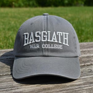 May include: A gray baseball cap with the text "BASGLATH WAR COLLEGE" embroidered in white. The cap has a curved brim and a button on top. The hat is made of a soft fabric and is suitable for casual wear.