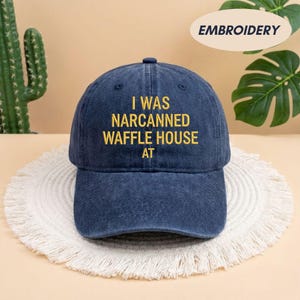 May include: A navy blue baseball cap with the embroidered text "I WAS NARCANNNED WAFFLE HOUSE AT" in yellow. The cap is sitting on a white, fringed placemat. The word "EMBROIDERY" is in a white oval in the background.