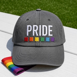 May include: A charcoal gray baseball cap featuring the word PRIDE embroidered in white capital letters. Below the text are six small, square embroidered patches in red, orange, yellow, green, blue, and purple. A folded rainbow flag is visible.