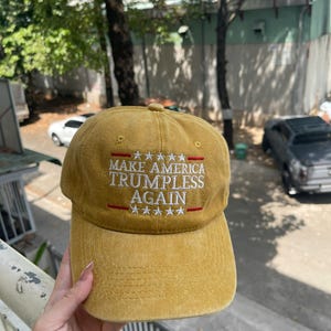 May include: Mustard-colored baseball cap with the text "MAKE AMERICA TRUMPLESS AGAIN" in white, with red accents and star graphics. The cap has a curved brim and a classic six-panel design.