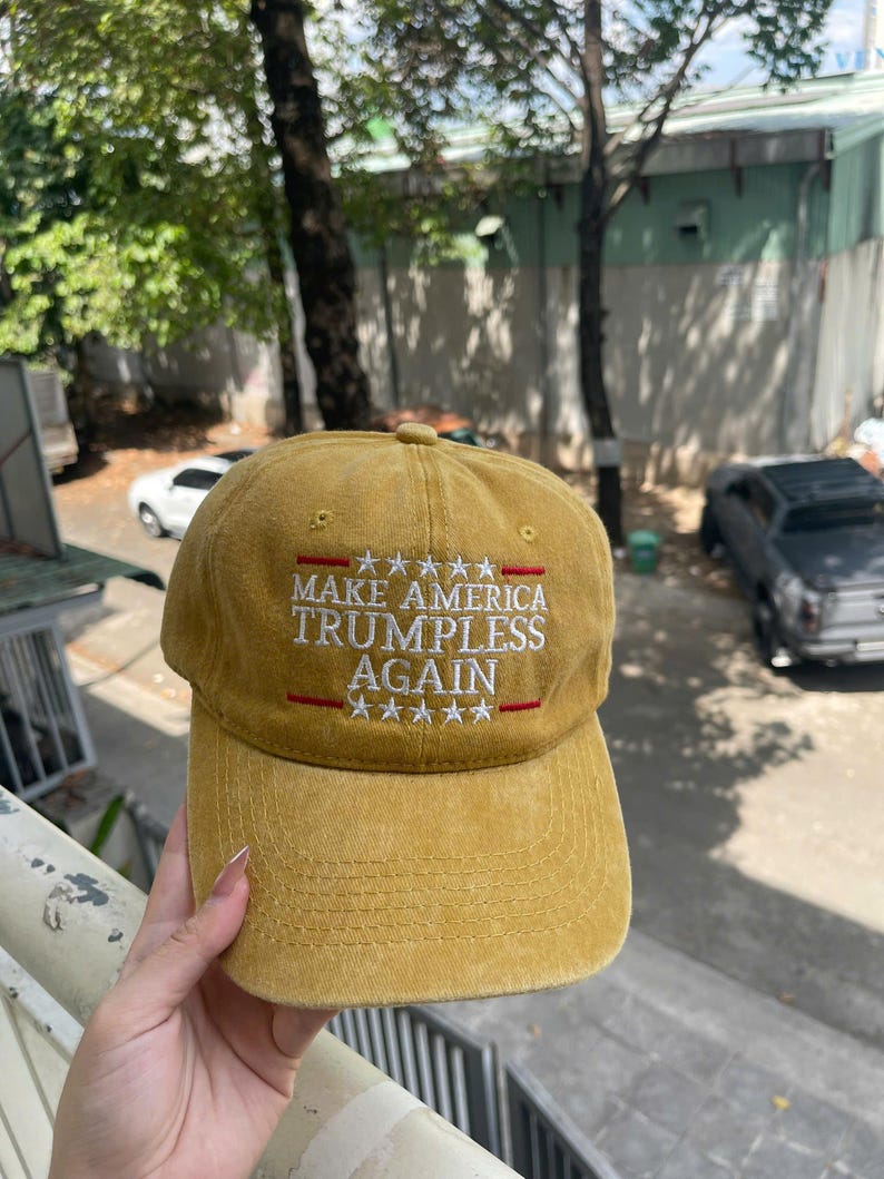 May include: Mustard-colored baseball cap with the text "MAKE AMERICA TRUMPLESS AGAIN" in white, with red accents and star graphics. The cap has a curved brim and a classic six-panel design.