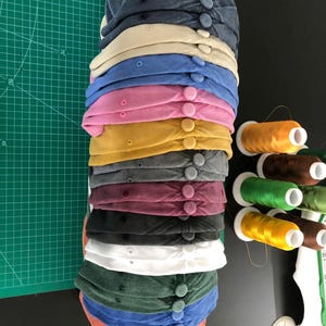 May include: A stack of baseball caps in various colors, including navy, tan, blue, pink, and yellow. Each cap has a button on top. Spools of thread in various colors are visible on the right.