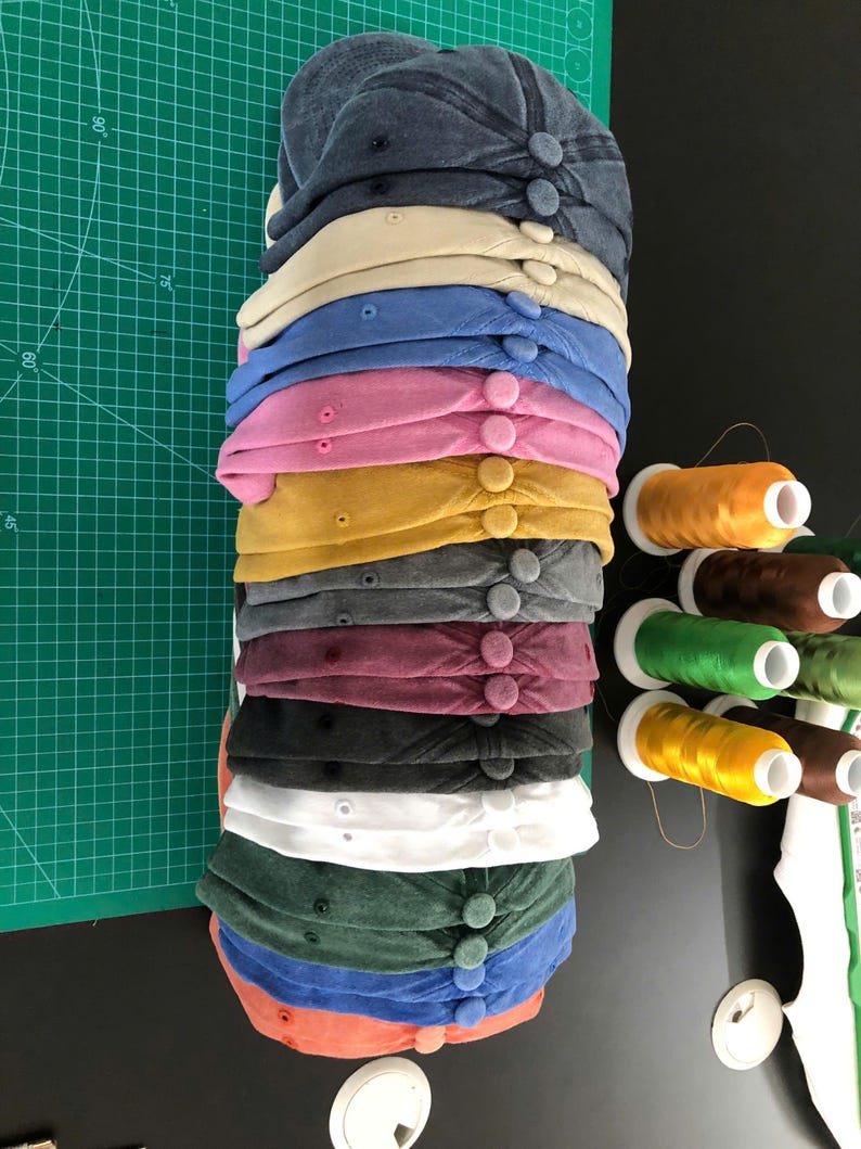 May include: A stack of baseball caps in various colors, including navy, tan, blue, pink, and yellow. Each cap has a button on top. Spools of thread in various colors are visible on the right.