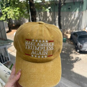 May include: Mustard yellow baseball cap with the text "MAKE AMERICA TRUMPLESS AGAIN" in white, with red accents. The cap has a curved brim and a classic six-panel design.