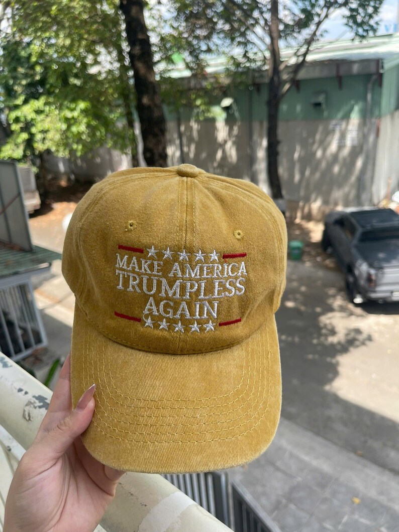 May include: Mustard yellow baseball cap with the text "MAKE AMERICA TRUMPLESS AGAIN" in white, with red accents. The cap has a curved brim and a classic six-panel design.
