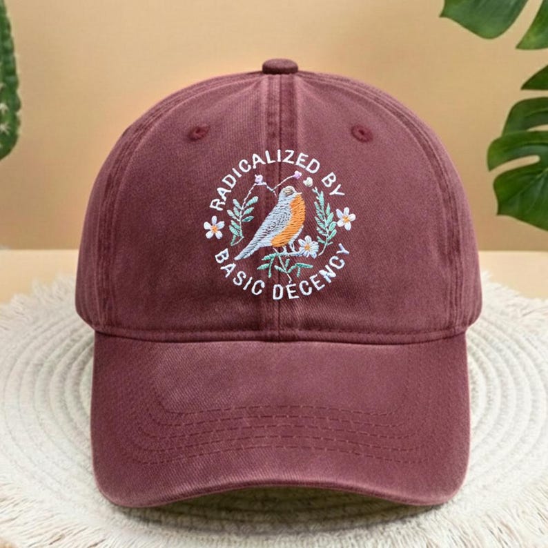 May include: A maroon baseball cap with an embroidered bird and floral design. The text "RADICALIZED BY BASIC DECENCY" is embroidered in a circle. The cap has a curved peak and an adjustable strap for a comfortable fit.