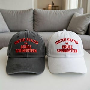 May include: Two baseball caps, one gray and one white, are displayed side-by-side. Both caps feature the text "UNITED STATES vs. BRUCE SPRINGSTEEN" embroidered in red. The caps are sitting on a white surface.