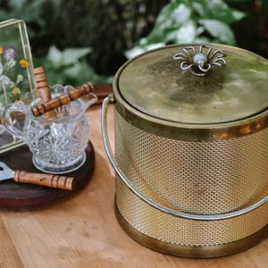 May include: A cylindrical gold-toned metal ice bucket with a textured woven pattern, a matching lid featuring a flower-shaped handle, and a curved metal swing handle. Beside it sits a clear glass creamer, a bottle opener, and a pressed flower frame.