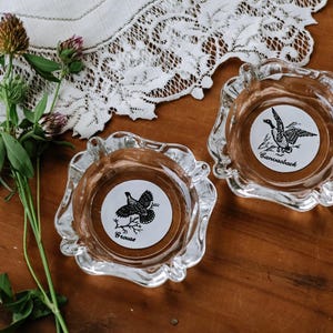 May include: Two clear glass ashtrays with scalloped edges. One ashtray features a black and white graphic of a grouse with the text "Grouse" below it. The other ashtray features a black and white graphic of a canvasback duck with the text "Canvasback" below it.