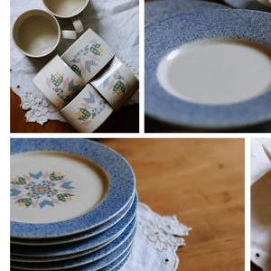 May include: A collection of vintage-style dinnerware featuring a blue speckled rim. The set includes stacked plates, mugs, and a plate with a floral quilt pattern in the center. The plates and mugs are arranged on a white lace doily.