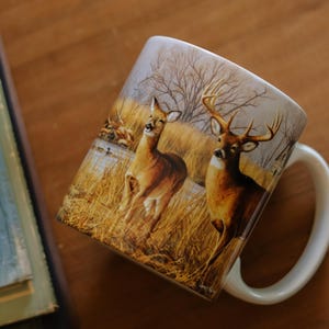 May include: A white ceramic mug featuring a detailed wrap-around illustration of two deer in a grassy field. One deer is a buck with large antlers, and the other is a doe. The scene includes tall golden grass, bare trees, and a calm body of water.