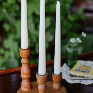 May include: A wooden candle holder with a circular base and three vertical pillars of varying heights, each topped with a white taper candle. The wood features a visible grain pattern and a smooth, polished finish, holding the candles upright.