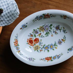 May include: An oval white ceramic serving bowl featuring a delicate floral pattern with orange, blue, yellow, and purple flowers and green leaves arranged in a wreath-like design around the interior base and along the inner rim of the bowl.