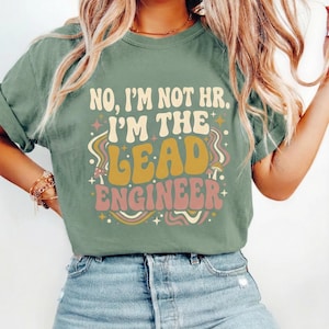 May include: A sage green t-shirt featuring the text "NO, I'M NOT HR. I'M THE LEAD ENGINEER" in a retro style. The design incorporates stars, mushrooms, and wavy lines in yellow, pink, and cream. The shirt is worn with blue denim jeans.