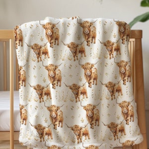 May include: A cream-colored baby blanket with a repeating pattern of brown Highland cows and gold floral accents. The blanket is draped over a wooden crib, showcasing its soft, plush texture. The blanket is designed for a nursery.