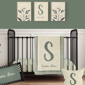 May include: A nursery set featuring a black crib with a green and white gingham patterned sheet. The set includes a blanket, pillow, and a rectangular pillow, all with the letter "S" and the name "Sierra Remi". Three framed floral prints hang above the crib.