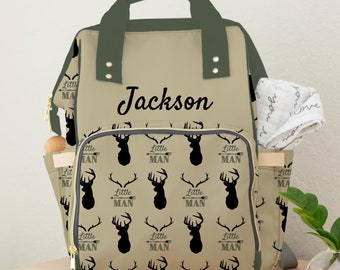 May include: A beige and green diaper bag with a black and white deer print. The bag has a zippered pocket on the front and a personalized name tag that says "Jackson".
