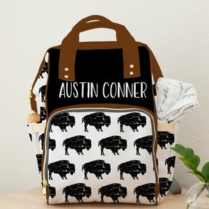 May include: A black and white diaper bag with a brown leather trim. The bag has a buffalo plaid pattern and the name "Austin Conner" is printed on the front.