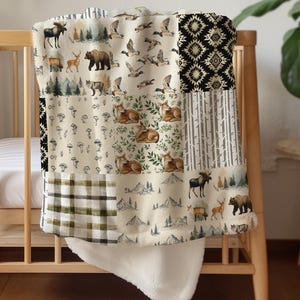 May include: A patchwork blanket draped over a wooden cot. The blanket features various nature-themed patterns, including woodland animals, trees, and geometric designs in beige, black, and green. The blanket has a soft, white, fluffy underside.