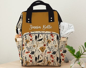 May include: A mustard yellow diaper bag with black straps and a floral print. The bag has a zippered pocket on the front and a name tag that reads "Zinnia Belle".