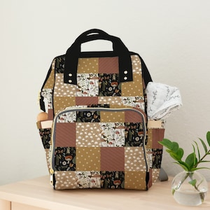 May include: A brown and black patchwork diaper bag with gold accents and a zipped pocket. The bag has a quilted design with a floral and mushroom pattern.