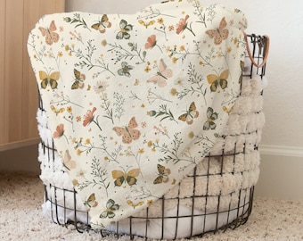 May include: A cream-colored throw blanket with a floral and butterfly pattern in shades of yellow, orange, and green. The blanket is folded and placed in a wire basket.