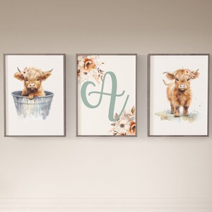 Puede incluir: Tres impresiones enmarcadas con ilustraciones de acuarela de una vaca de las tierras altas. La primera impresi&oacute;n muestra una vaca sentada en un cubo, la segunda impresi&oacute;n muestra la letra "A" con acentos florales, y la tercera impresi&oacute;n muestra una vaca de pie sobre un fondo de acuarela.