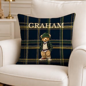 May include: A square throw pillow featuring a dark blue, green, and yellow plaid pattern. The name GRAHAM is printed in light-colored capital letters above a graphic of a teddy bear wearing a green bucket hat, a plaid jacket, and tan pants.