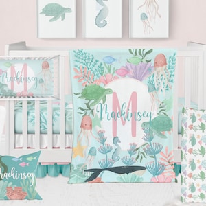 May include: A baby crib bedding set with a blue and green ocean theme. The set includes a crib bumper, a fitted sheet, a blanket, and a pillow. The blanket features a large pink letter M with the name Mackinsey written in cursive. The crib bumper and fitted sheet feature a repeating pattern of sea creatures and coral.