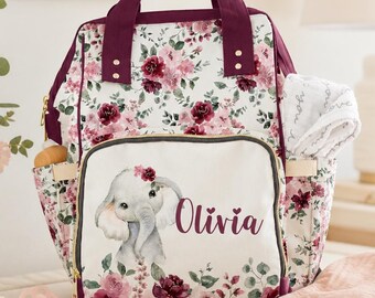 May include: A diaper backpack featuring a white background with a floral pattern in shades of burgundy and pink. The front pocket displays a cartoon elephant illustration and the name Olivia in script. It has dark burgundy handles and trim.