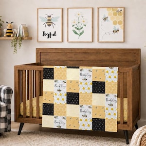 May include: A patchwork baby blanket featuring a repeating pattern of yellow honeycombs, black and yellow bees, and white fabric squares with the name Berkley printed in a black script font, draped over a wooden crib in a nursery setting.