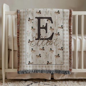 May include: A woven throw blanket featuring a repeating pattern of mallard ducks and reeds on a beige background. The center displays a large dark brown letter E with the name Elliot written in cursive script below it. The edges have a fringe.